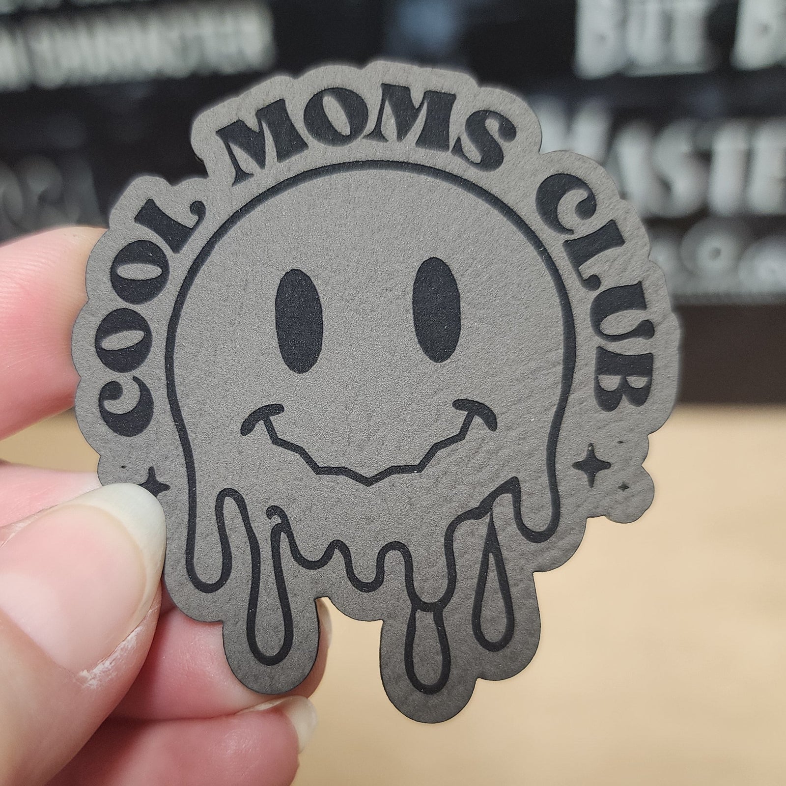 Patch with 'Cool Moms Club' design held in a hand against a blurred background