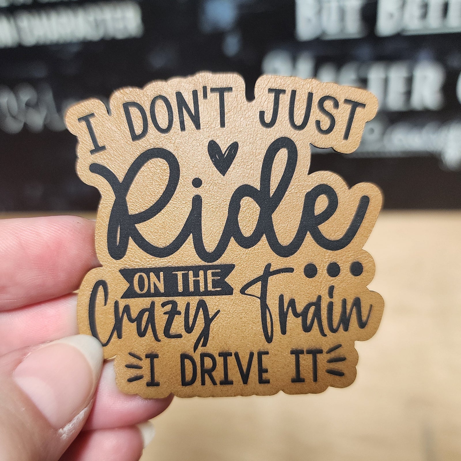 Hand holding a wooden sign with text 'I don't just ride on the crazy train, I drive it' against a blurred background.