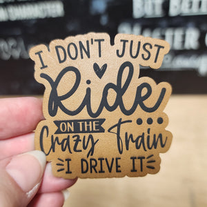 Hand holding a wooden sign with text 'I don't just ride on the crazy train, I drive it' against a blurred background.