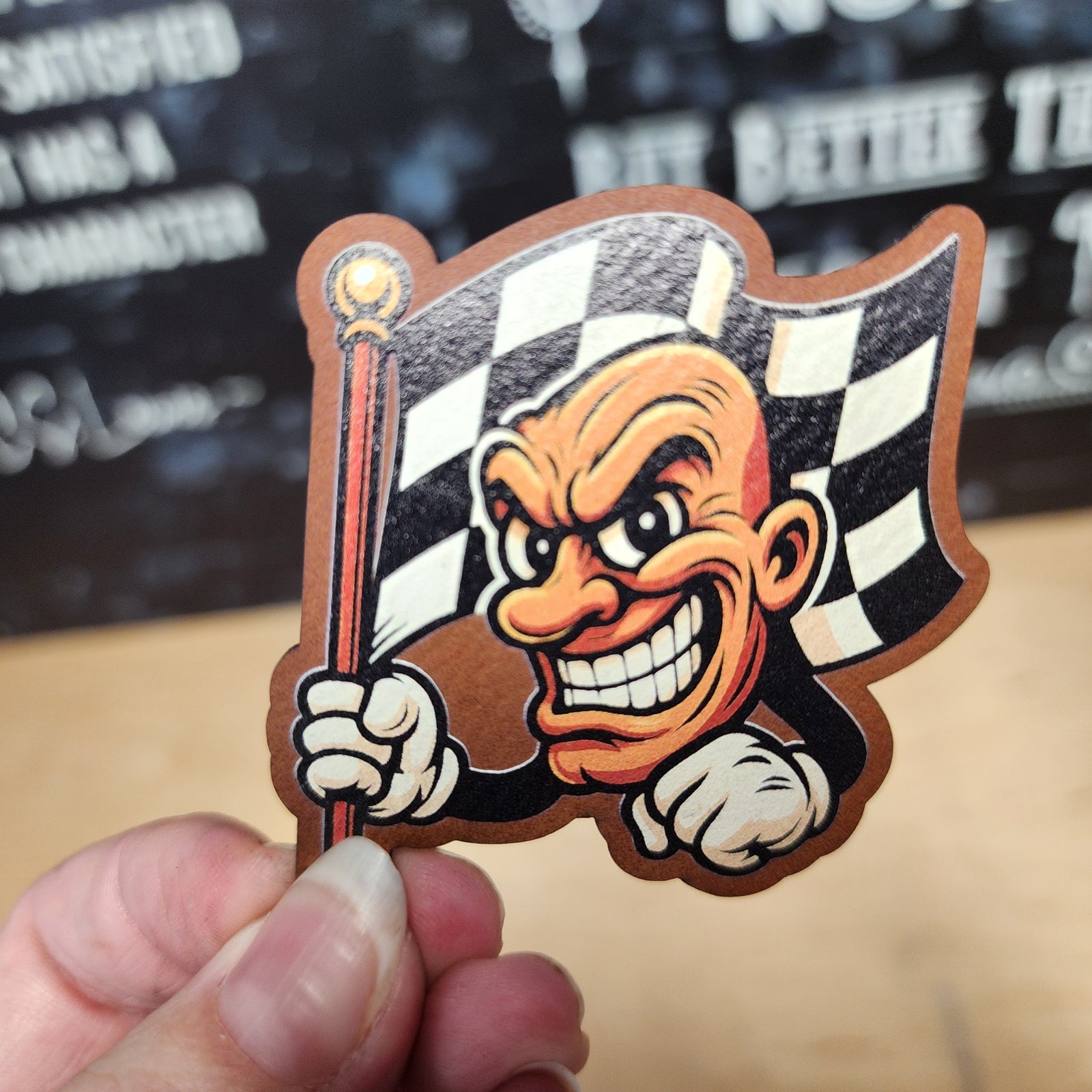 Racing mascot hat patch with a smiling character holding a checkered flag, perfect for motorsport enthusiasts.