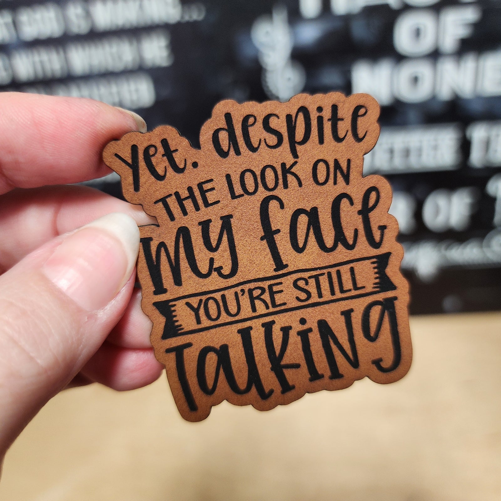 Humorous leatherette hat patch saying, 'Yet despite the look on my face, you’re still talking' for stylish accessories