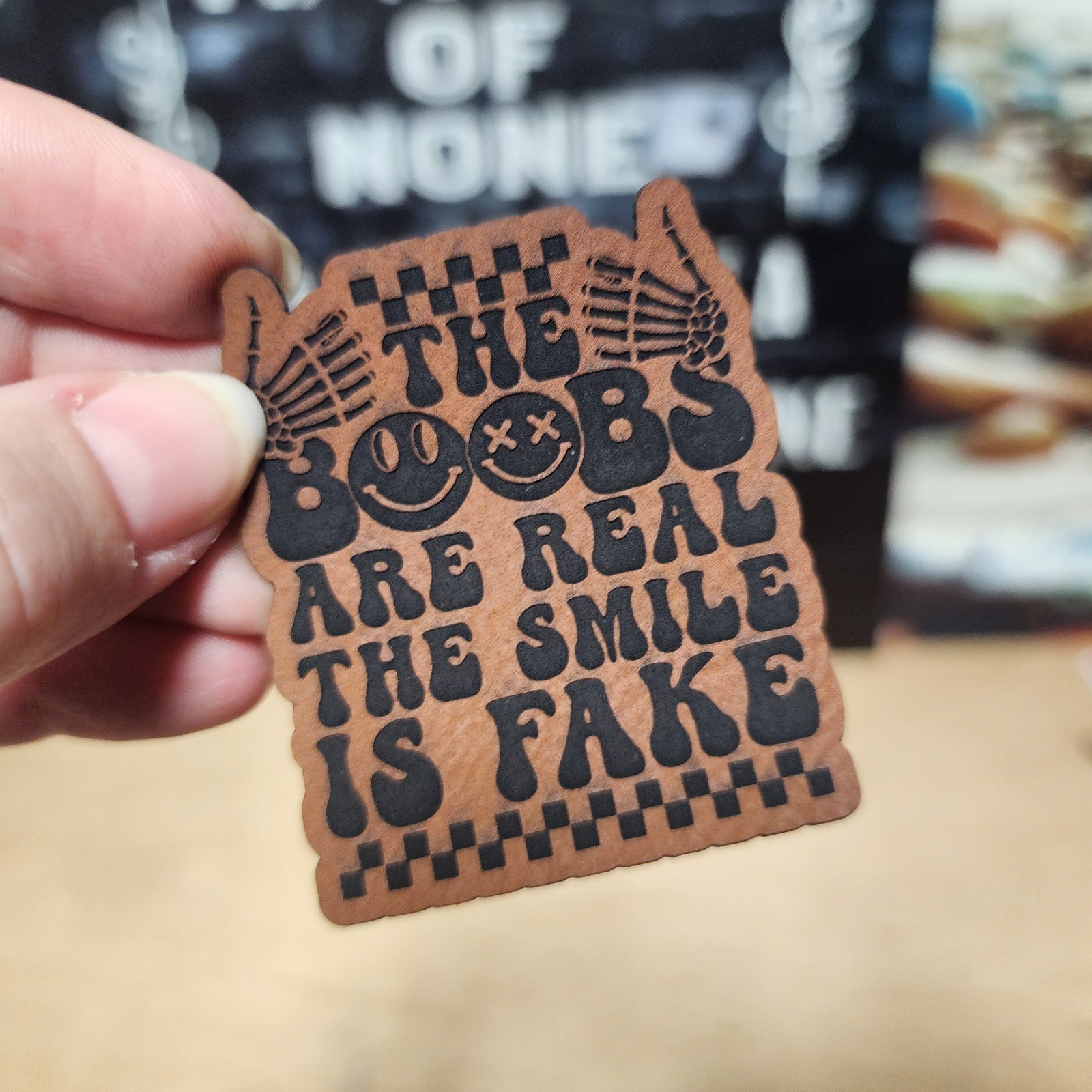 Humorous leatherette hat patch featuring the phrase 'The Boobs Are Real The Smile Is Fake', perfect for quirky accessories.