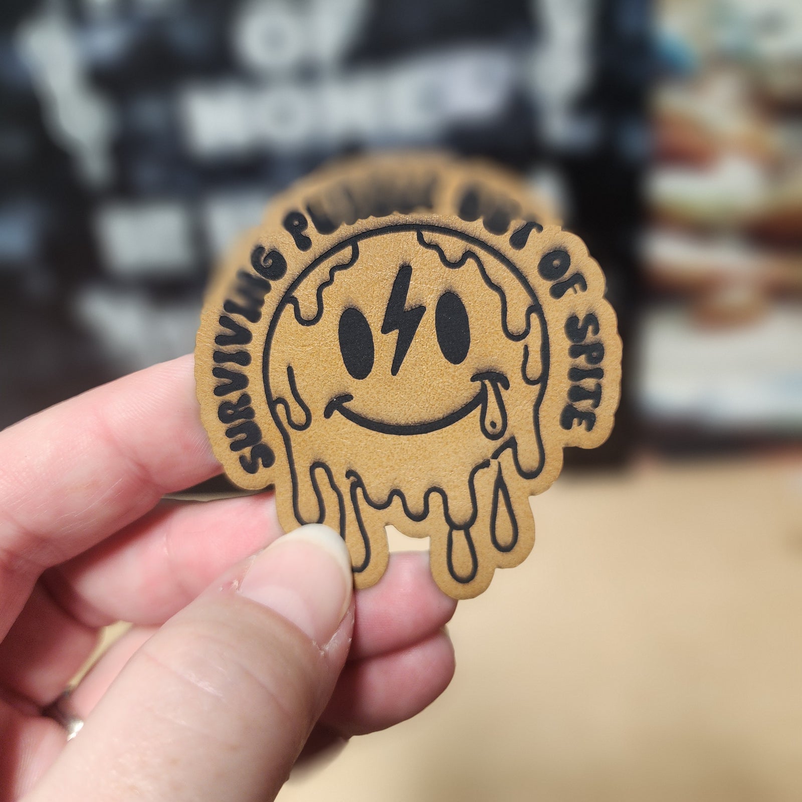 Funny leatherette hat patch featuring a smiling, melting smiley face with 'Surviving Planet Out of Spite' text.