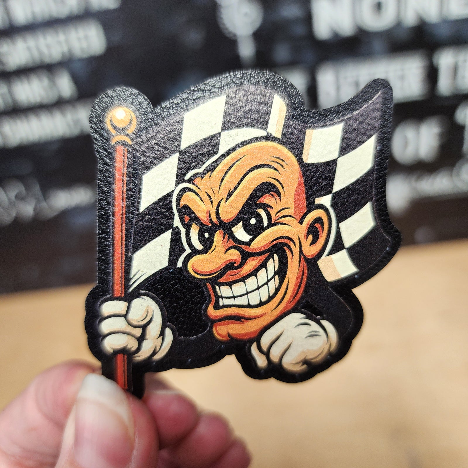Racing-themed leatherette hat patch featuring a grinning checkered flag mascot for motorsports enthusiasts.