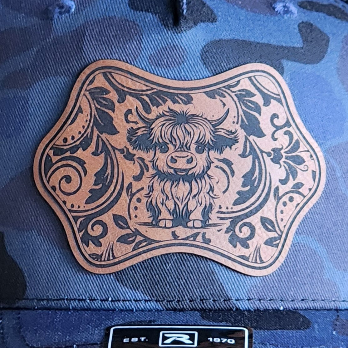 Cute Highland Cow leatherette hat patch on camo cap, perfect for animal lovers and stylish headwear enthusiasts.