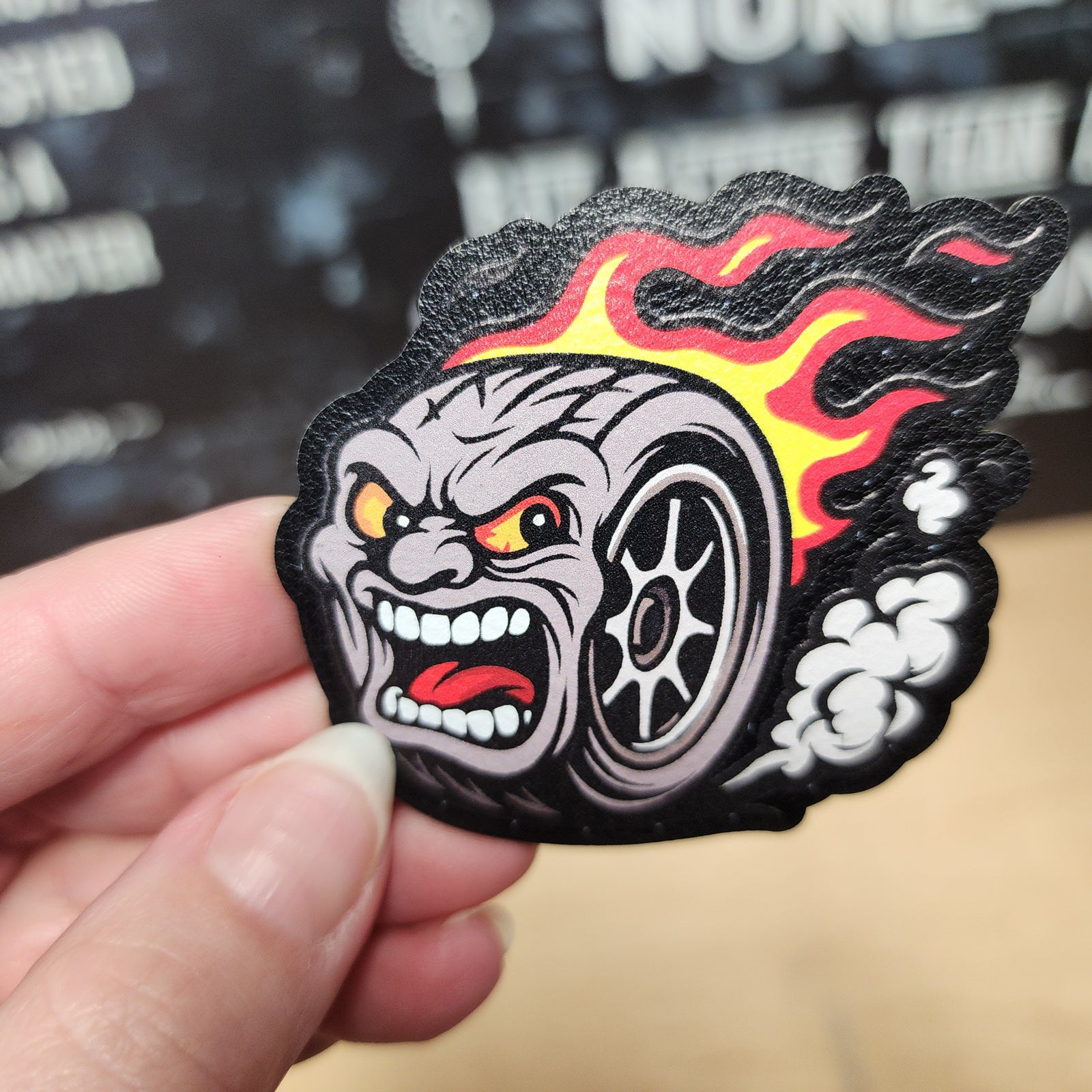 Cool racing tire leatherette patch with angry cartoon face and flames, perfect for automotive enthusiasts.