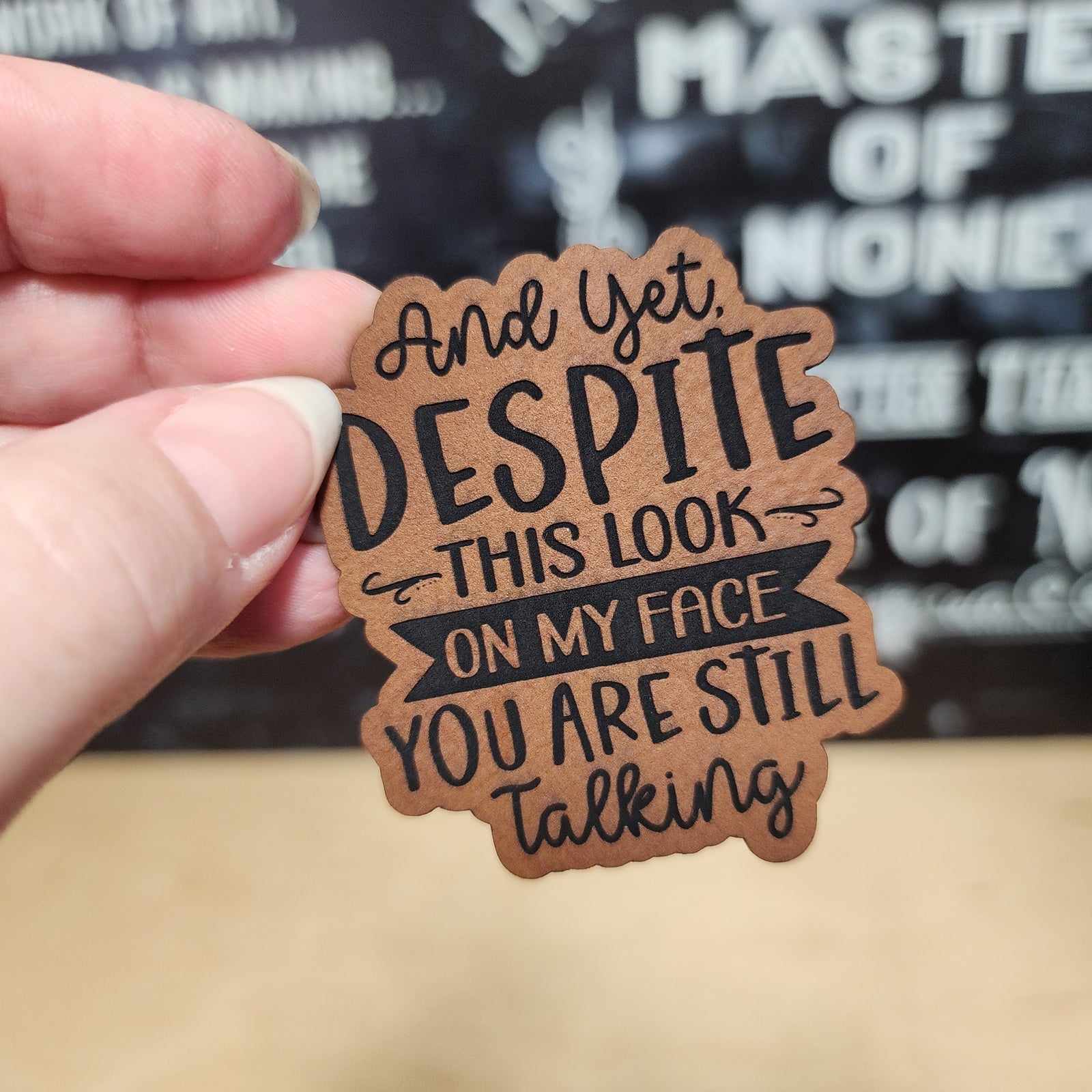 Funny leatherette hat patch with saying: 'And yet, despite this look on my face, you are still talking'.