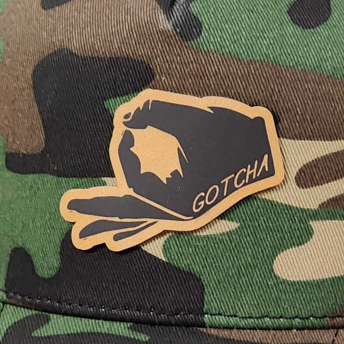 Gotcha leatherette hat patch with hand gesture design on camouflage background for trendy outdoor gear enthusiasts.
