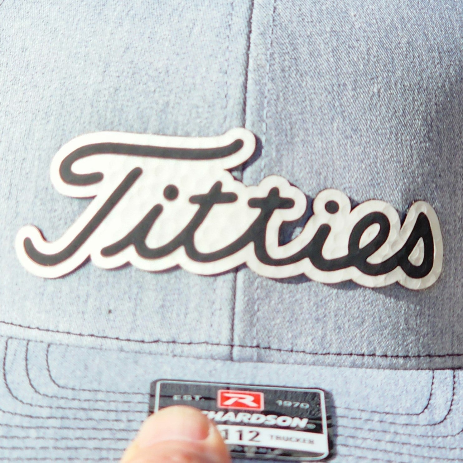Humorous leatherette hat patch reading 'Titties' on a stylish light blue cap for casual wear.
